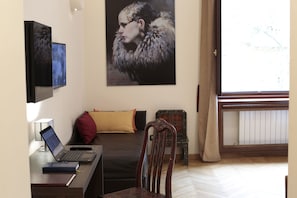 Een slaapkamer met een bed, een bureau met een laptop, een stoel, een bijzettafeltje, een decoratieve stoel, een schilderij en een raam met gordijnen.