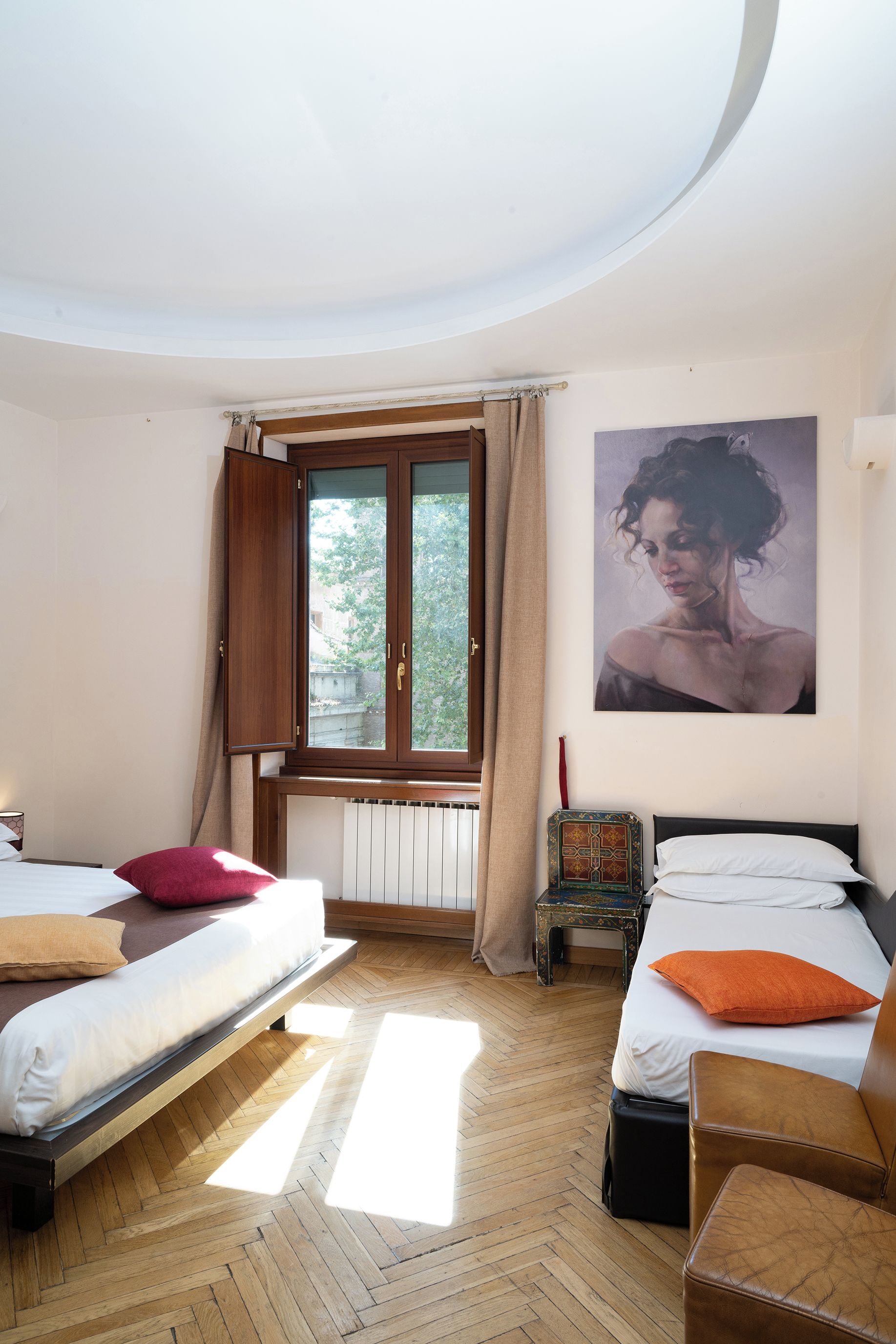 A hotel room with two beds, a window with curtains, a radiator, a small table, and a painting on the wall.