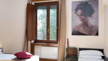 A hotel room with two beds, a window with curtains, a radiator, a small table, and a painting on the wall.