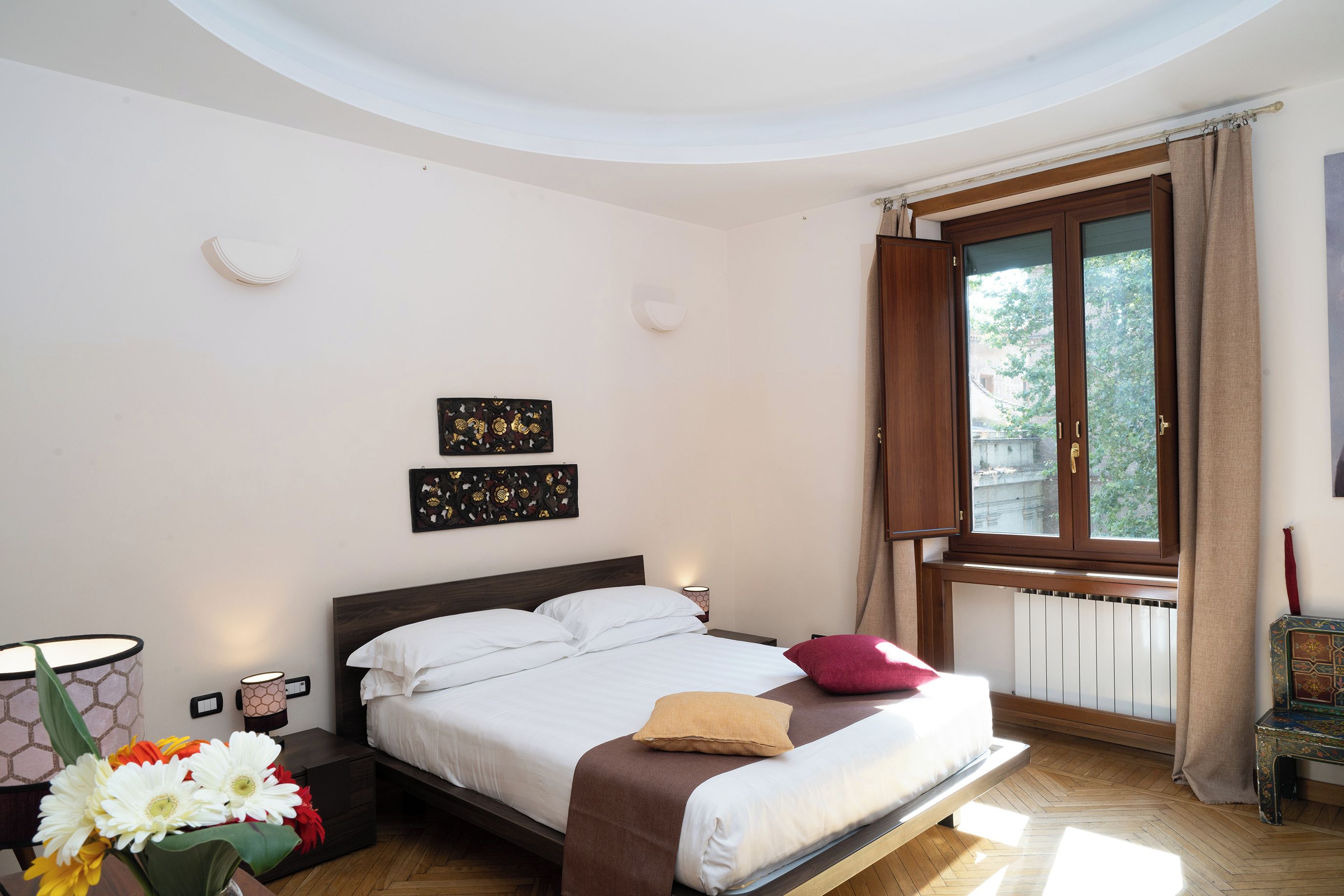 Een slaapkamer met een bed, nachtkastje, lamp, raam met gordijnen en uitzicht op het buiten.
