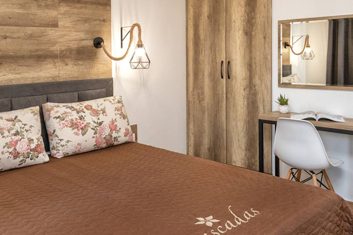 A bedroom with a wooden headboard, a bed with a brown quilt, two floral pillows, a desk with a chair, a potted plant, and a mirror on the wall.