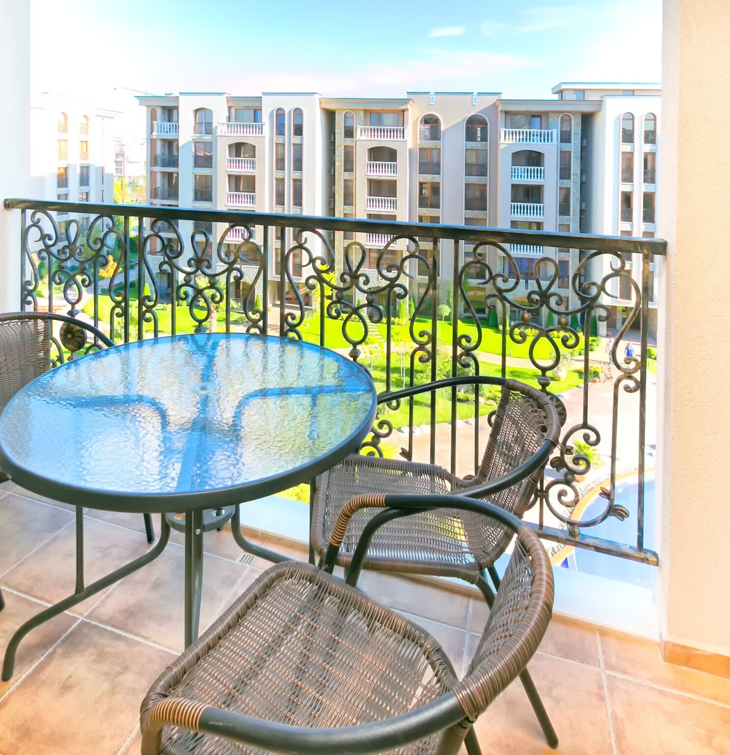 A balcony with a round glass table and wicker chairs, overlooking a well-maintained garden and a row of buildings.