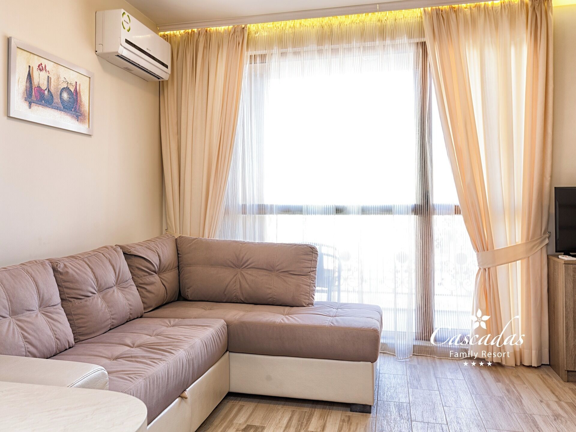 A modern living room with a sectional sofa, a flat-screen TV, and a large window with sheer curtains.