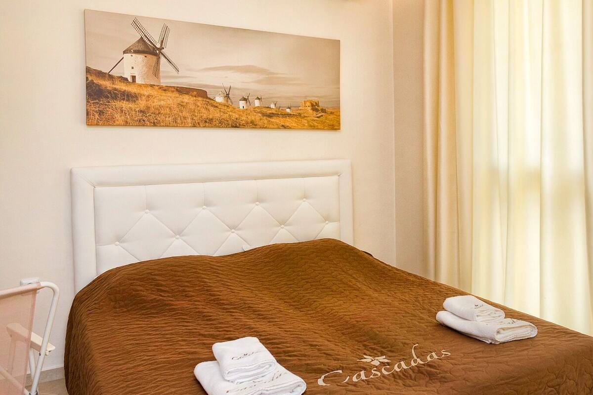 A hotel room with a tufted headboard, a brown bedspread, and a painting of windmills on the wall.