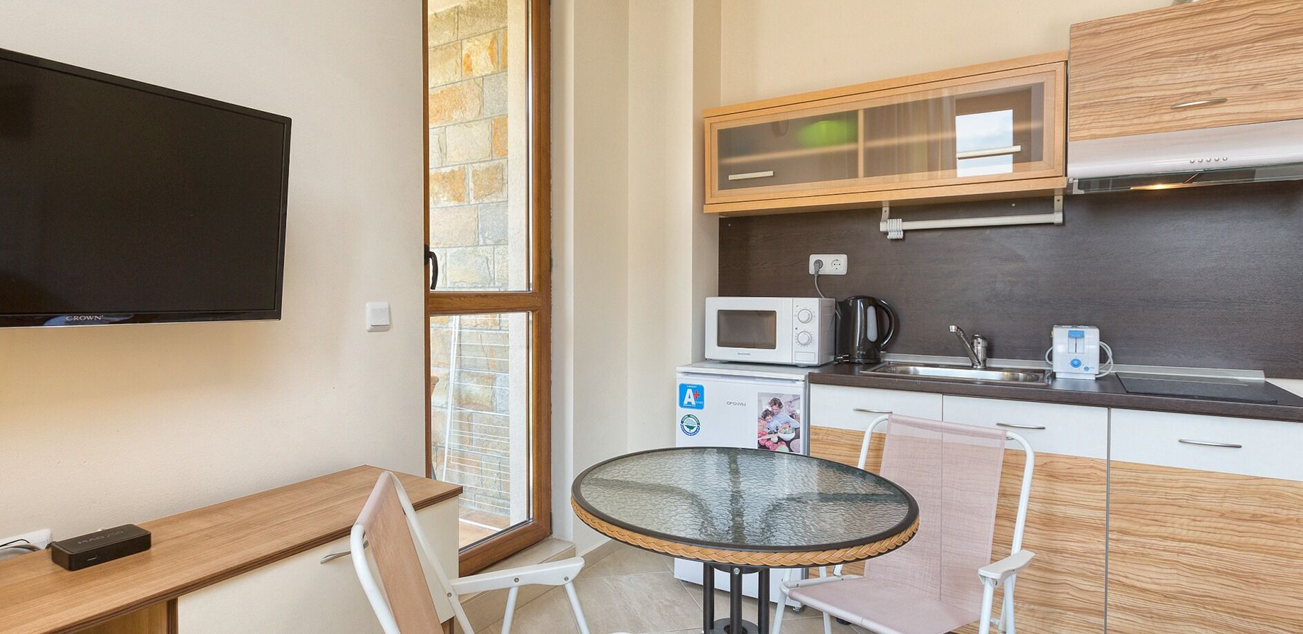 A compact kitchen with a microwave, kettle, and toaster, a small dining table with chairs, and a TV mounted on the wall.