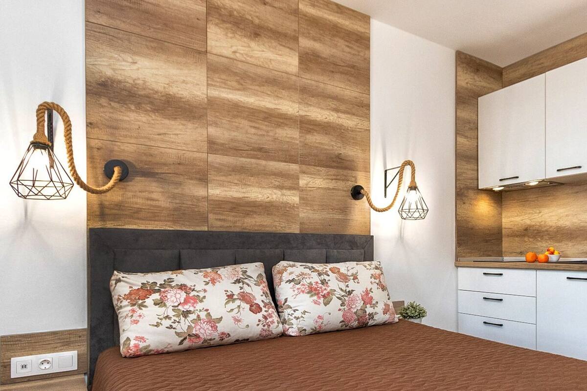 A modern bedroom with a large bed, two floral pillows, and a headboard against a wooden wall. Adjacent is a kitchen with white cabinets, a microwave, and a kettle on the counter.