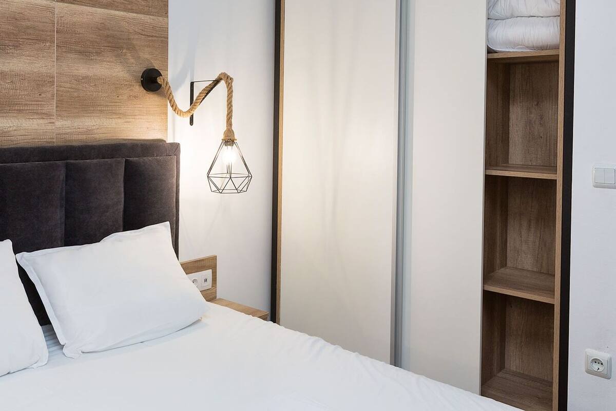 A neatly made bed with white linens and pillows, a modern headboard, a wall-mounted light fixture, and a wooden shelving unit.