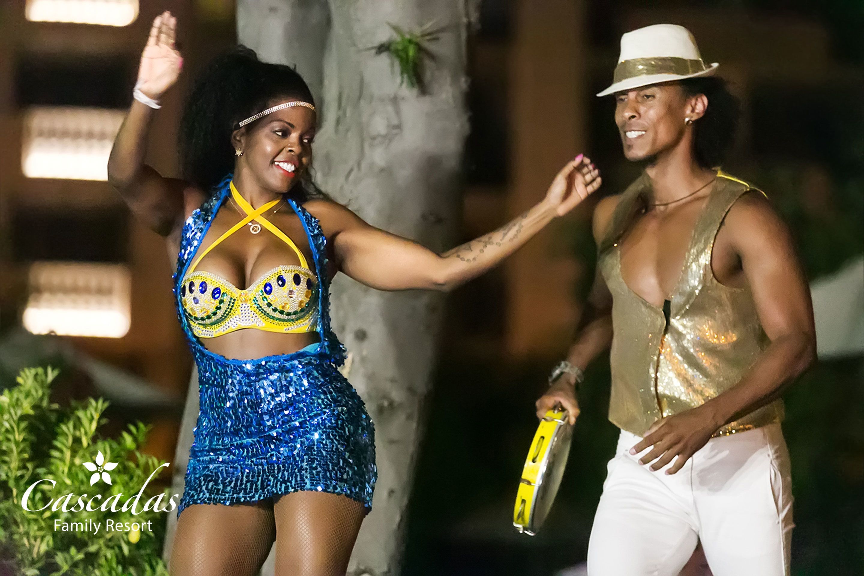 Dos personas bailando al aire libre de noche, una vestida con un traje azul lleno de lentejuelas y la otra con una blusa dorada y pantalones blancos.