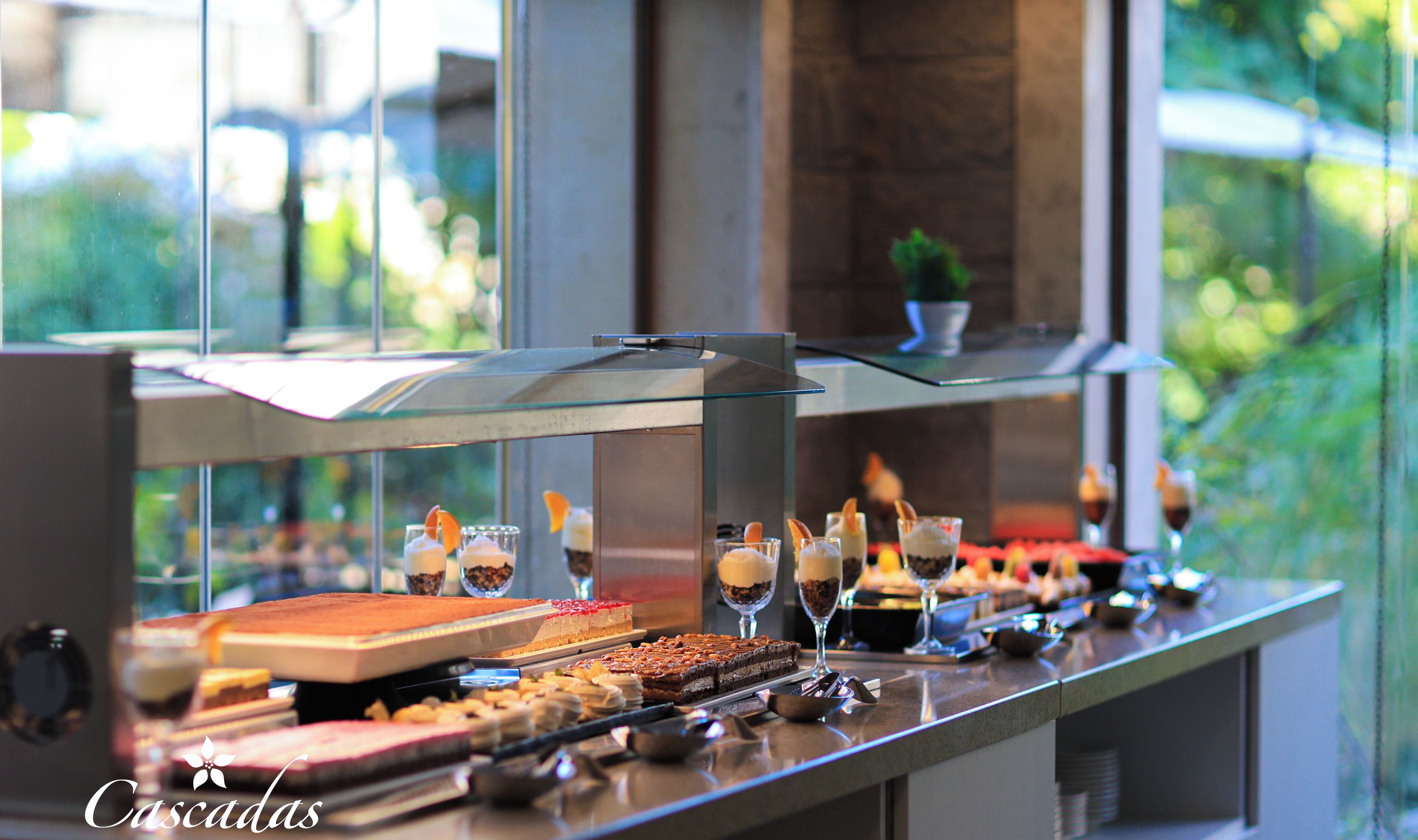 Mostrador de buffet con diversos postres y bebidas, una vitrina de vidrio y vistas a la vegetación exterior.