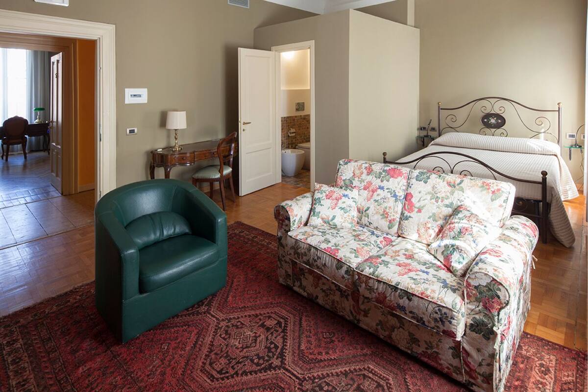 Un amplio salón con un sillón de cuero verde, un sofá floral y una alfombra roja estampada.