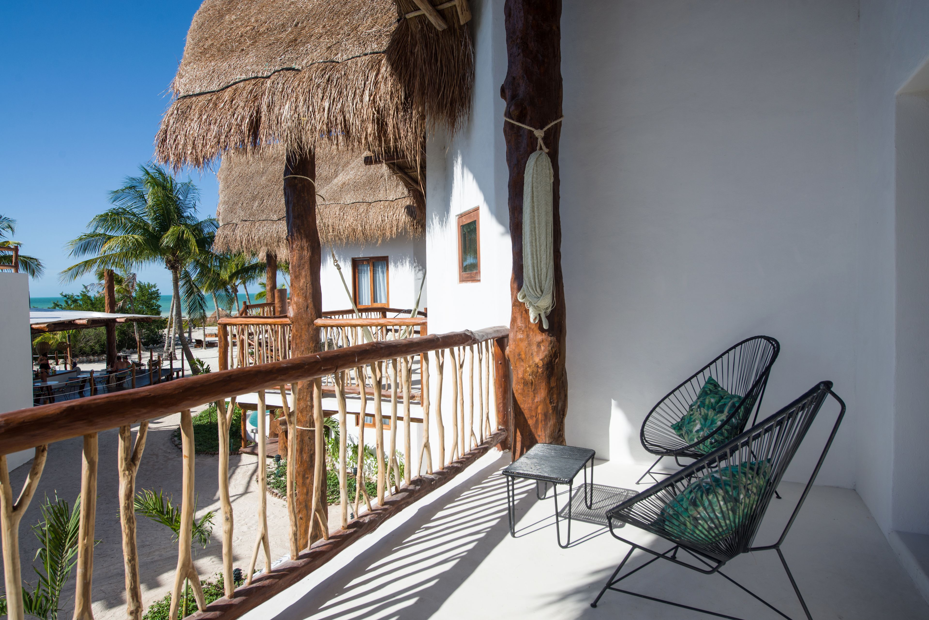 A balcony with a thatched roof, two wire mesh chairs, and a view of the beach and palm trees.