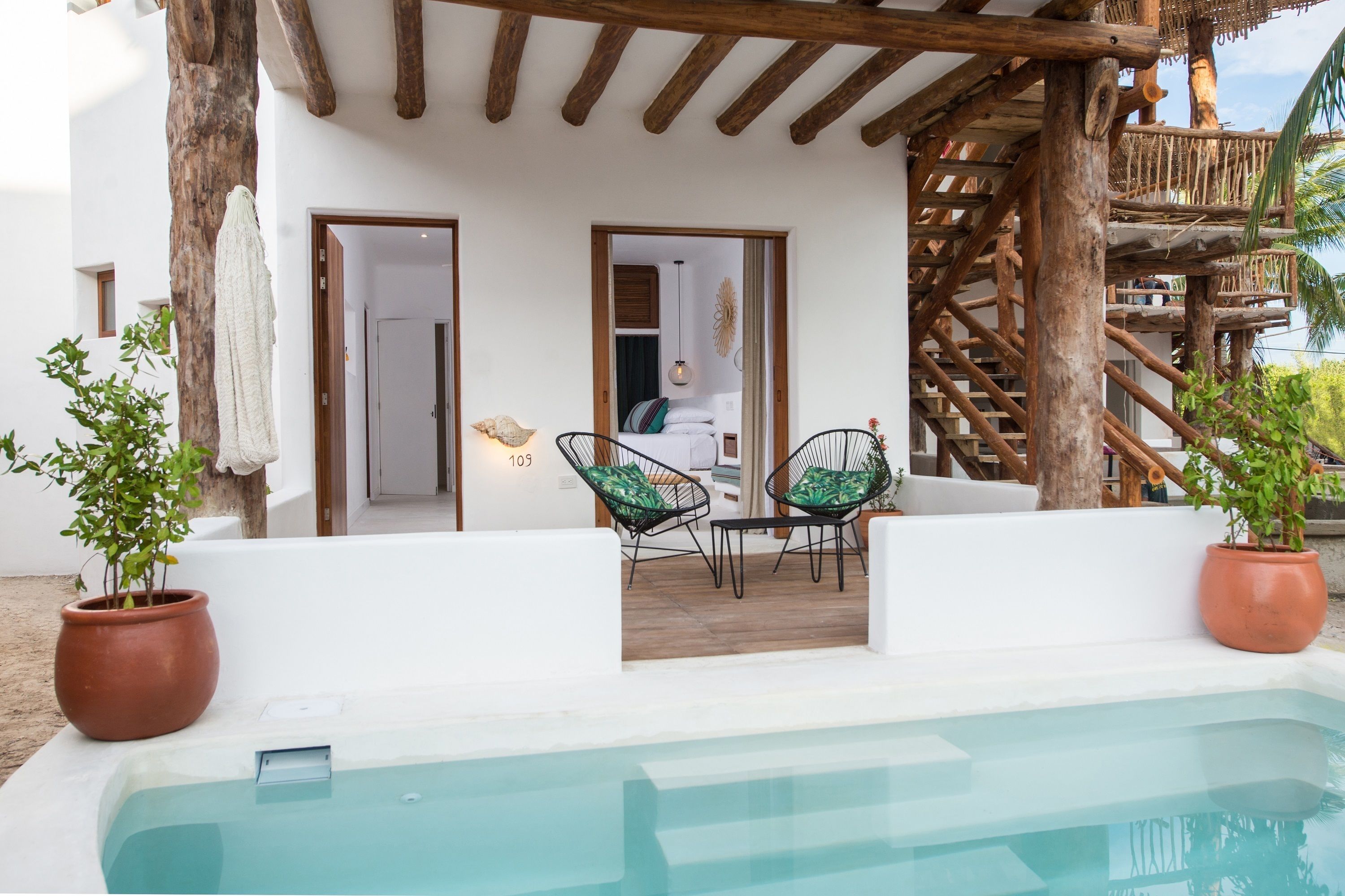 A poolside area with a wooden pergola, two green cushioned chairs, and a white wall with a door leading to a room.