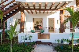 Uma área de estar coberta ao ar livre com uma pérgola de madeira, piscina e um edifício branco com porta e janelas.