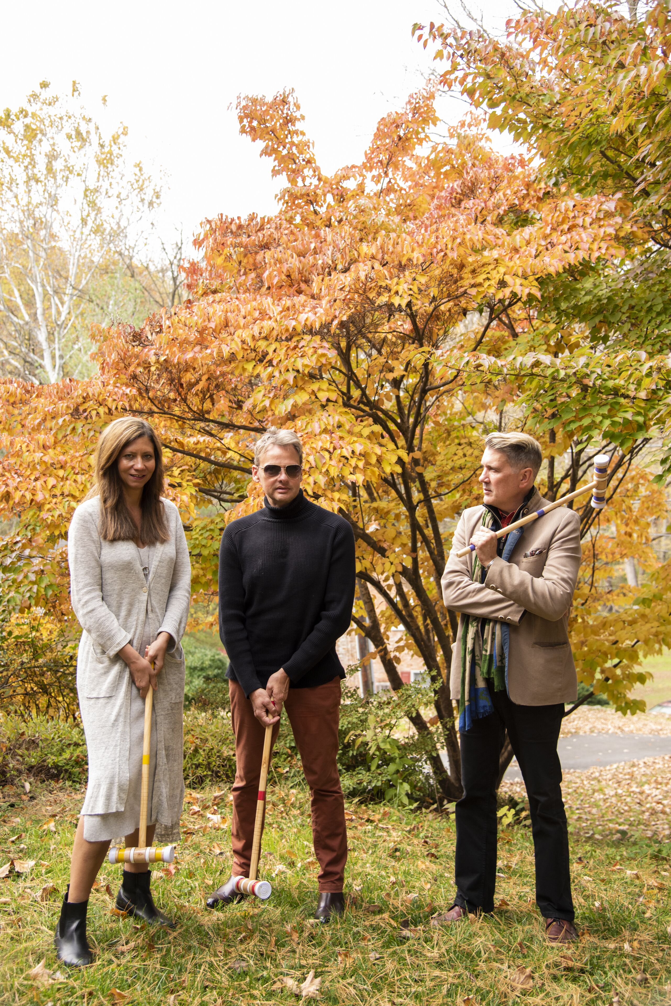 Three people standing outdoors with autumn leaves, holding sticks.