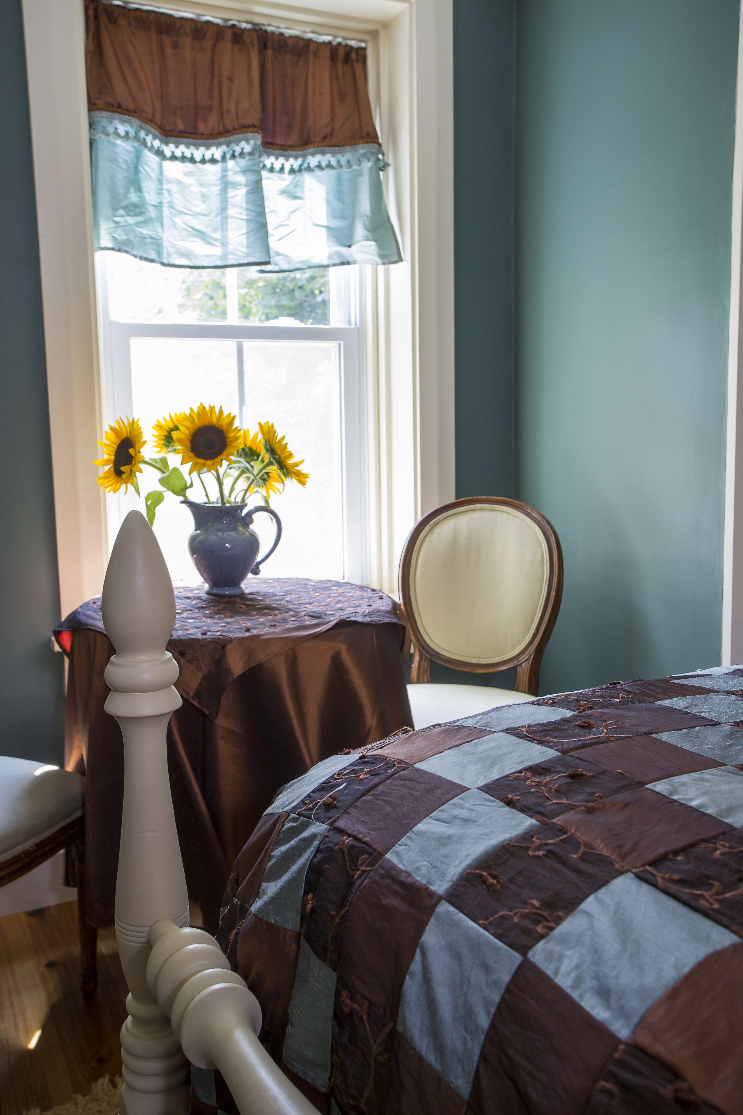 A bedroom with a bed, a chair, a table with a vase of sunflowers, and a window with curtains.