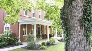 A brick building with a covered porch, potted plants, and a large tree. - The Hollinger House (Lancaster)