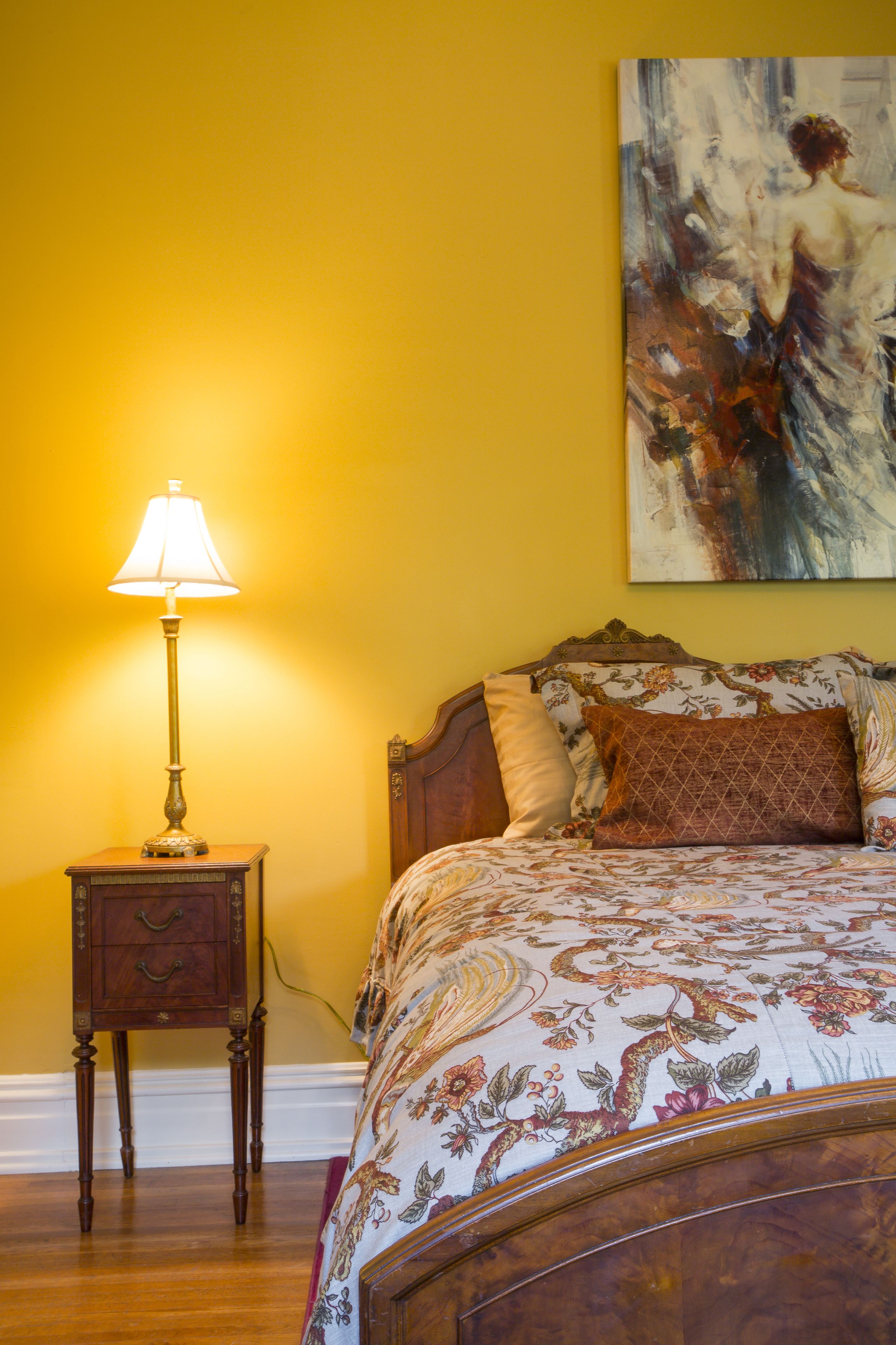 A bedroom with a wooden bed, floral bedding, a nightstand with a lamp, and a painting on the wall.