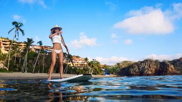 A person paddleboarding on a calm body of water with a clear sky and palm trees in the background.