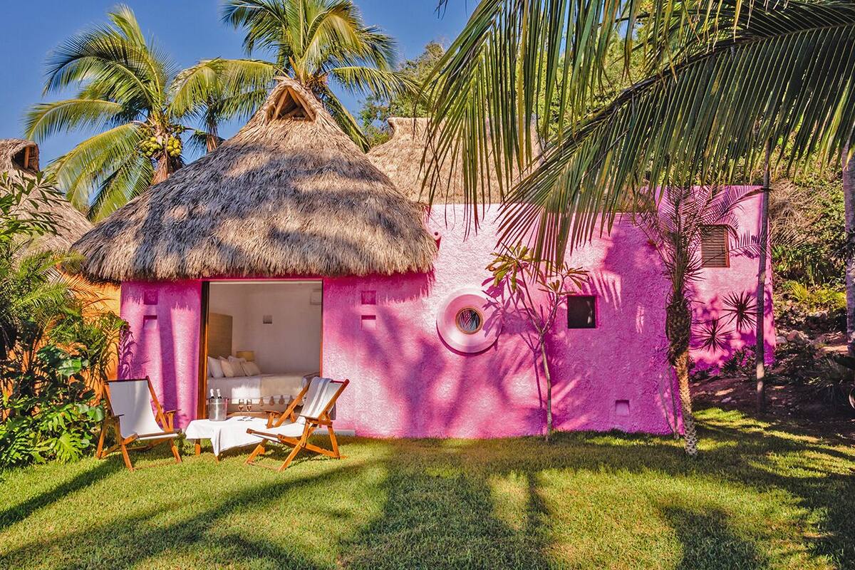 A pink building with a thatched roof, a small circular window, and a door leading to a room with a bed and a table.