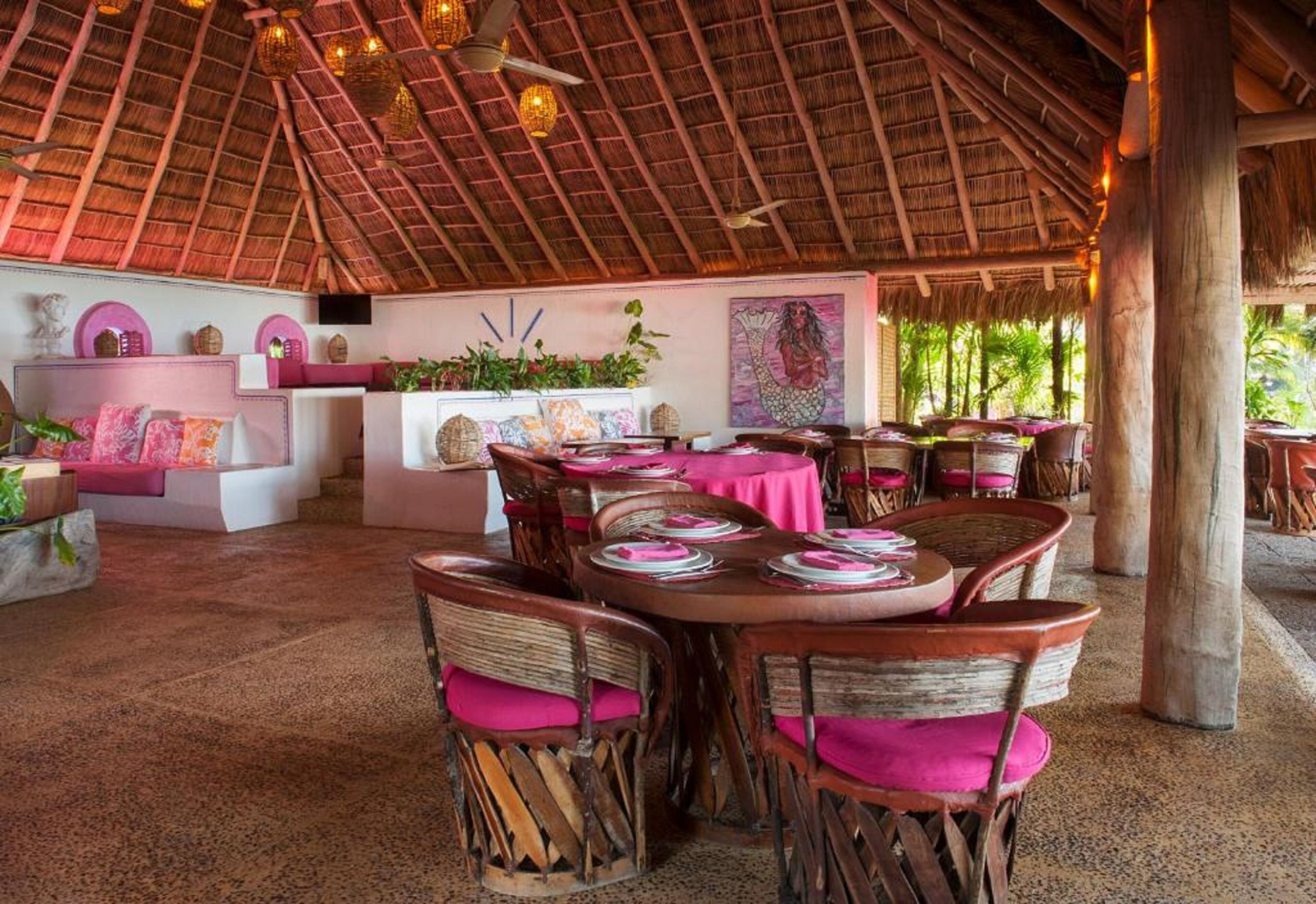 Un comedor interior con mesas y sillas de madera, techo de paja y un mural en la pared.