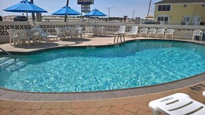 Une piscine dont le bord est recouvert de carrelage, entourée de chaises longues blanches et de parasols bleus.