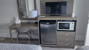 A hotel room with a wooden desk, a television, a microwave, a refrigerator, a chair, and a bed with patterned bedding. - Luna Mar Motel (Seaside Park)
