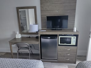A hotel room with a wooden desk, a television, a microwave, a refrigerator, a chair, and a bed with patterned bedding. - Luna Mar Motel (Seaside Park)