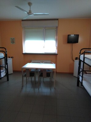 Un dormitorio con literas, una mesa con sillas, un ventilador de techo, una ventana con persianas y un televisor en la pared.