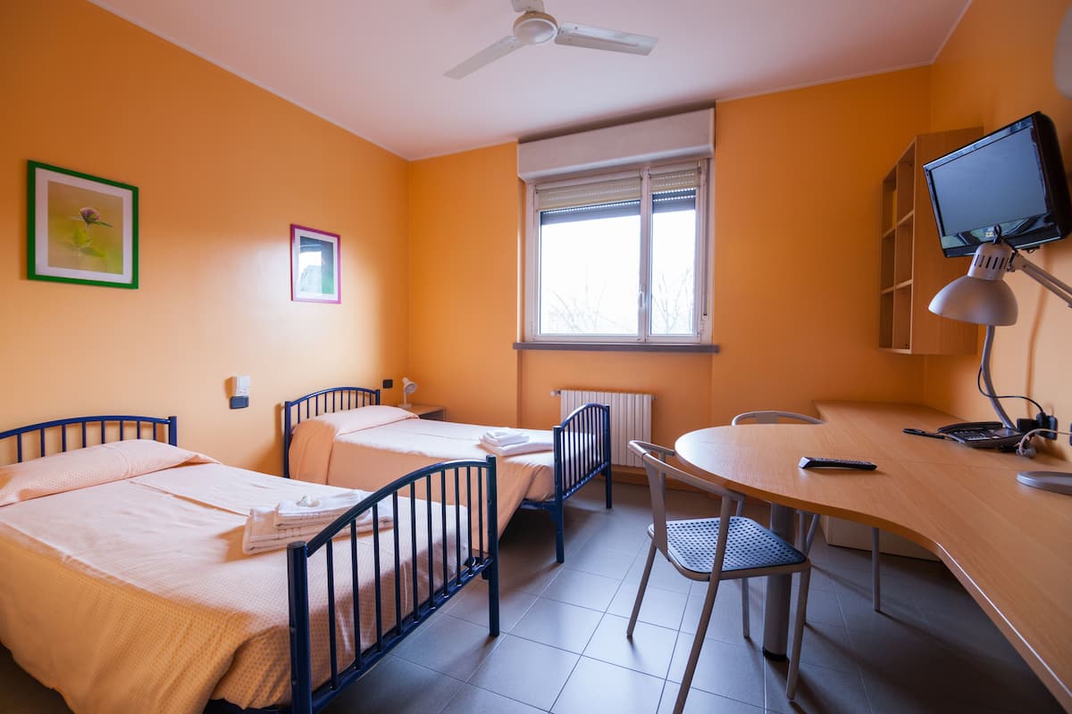 A room with two beds, a desk, a chair, a ceiling fan, a window, and a television.