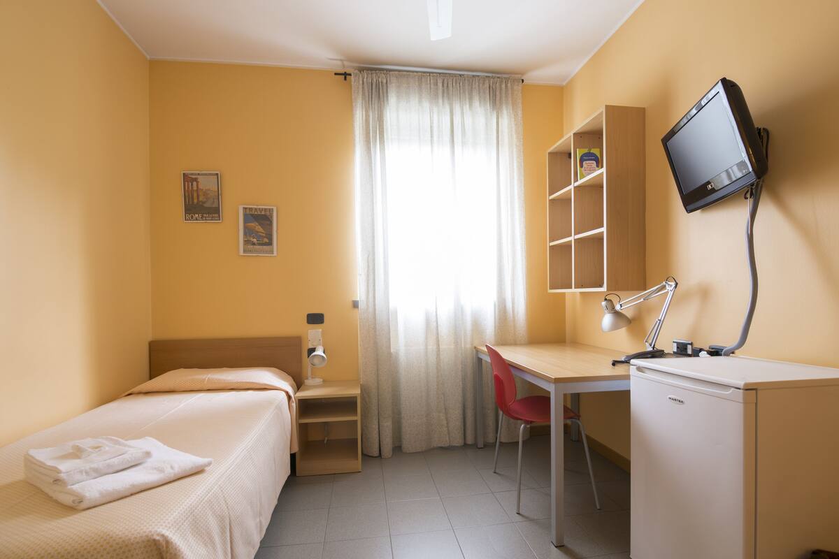 A hotel room with a bed, a desk with a chair, a television, and a window with curtains.
