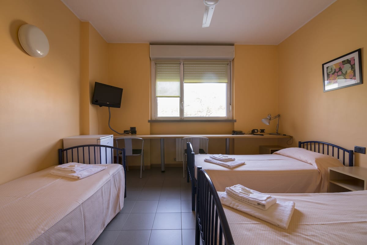 A hotel room with two single beds, a desk with a chair, a television mounted on the wall, and a window with blinds.