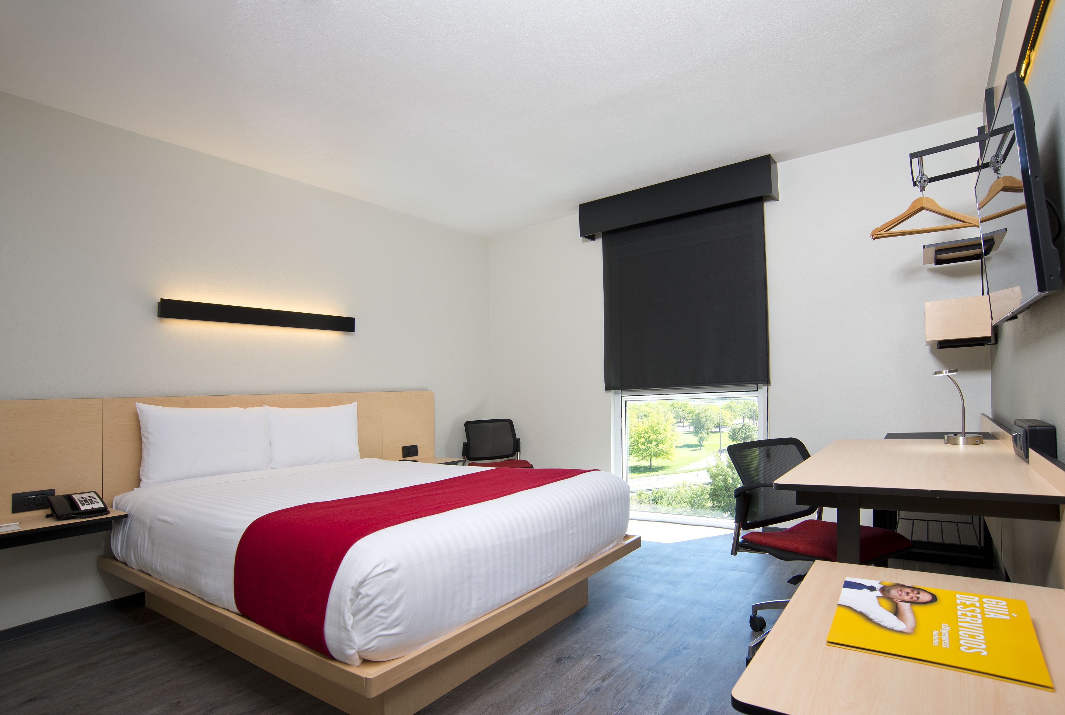 A modern hotel room with a large bed, a desk, a chair, a TV, and a window with a view of greenery.