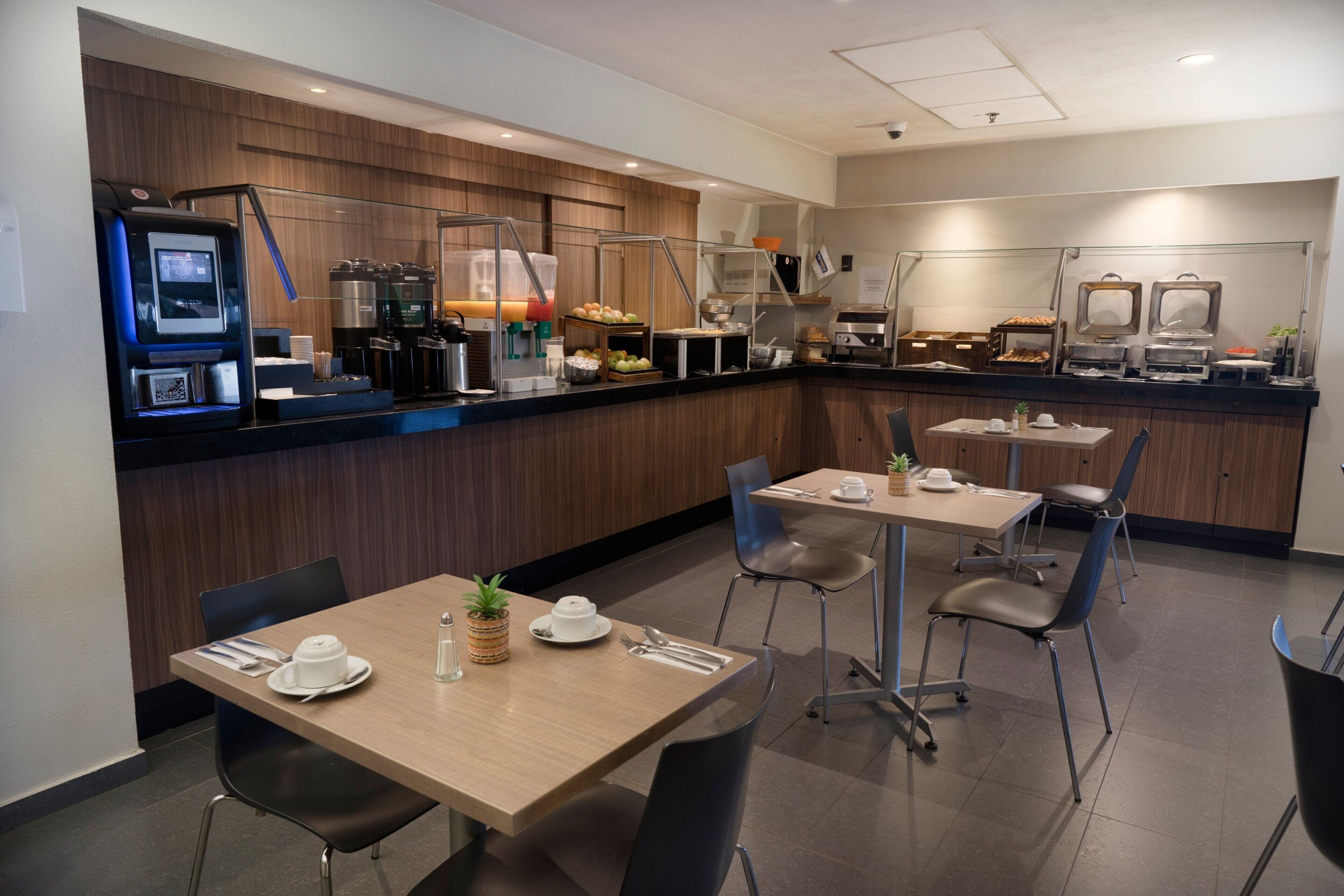A modern hotel breakfast area with self-service coffee machines, a buffet with various food items, and a seating area with tables and chairs.