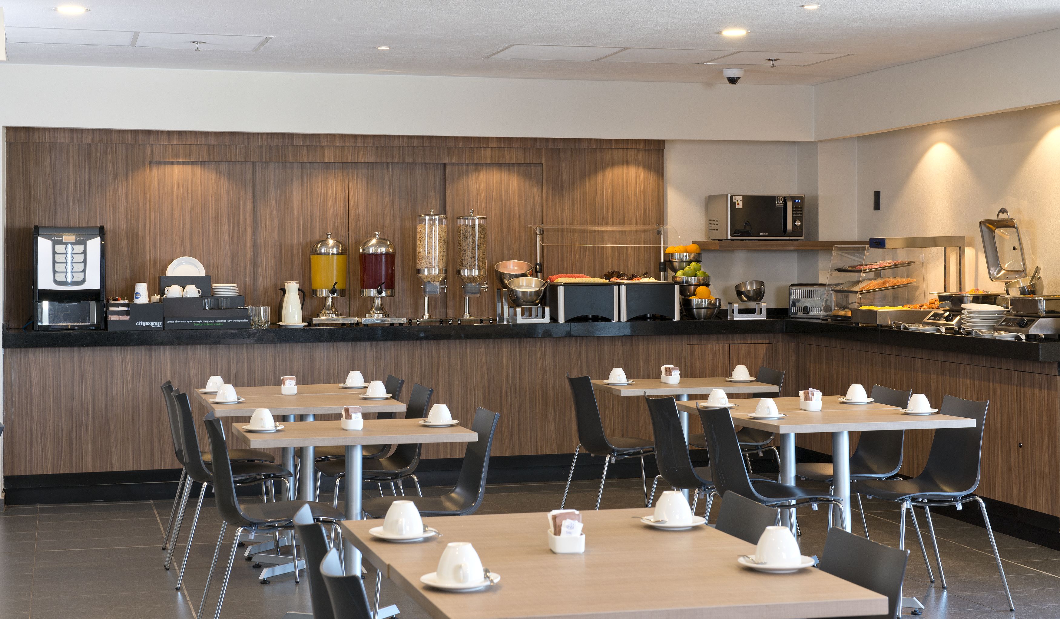 A modern breakfast bar with coffee machines, a microwave, and a variety of food options. Tables are set with white plates and cups.
