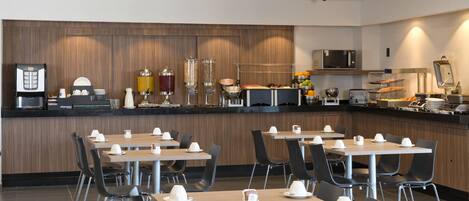 A modern breakfast bar with coffee machines, a microwave, and a variety of food options. Tables are set with white plates and cups.