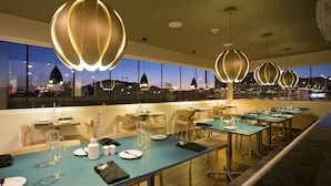 Een restaurant met gedekte tafels, grote hanglampen en uitzicht op de stad in de schemering.