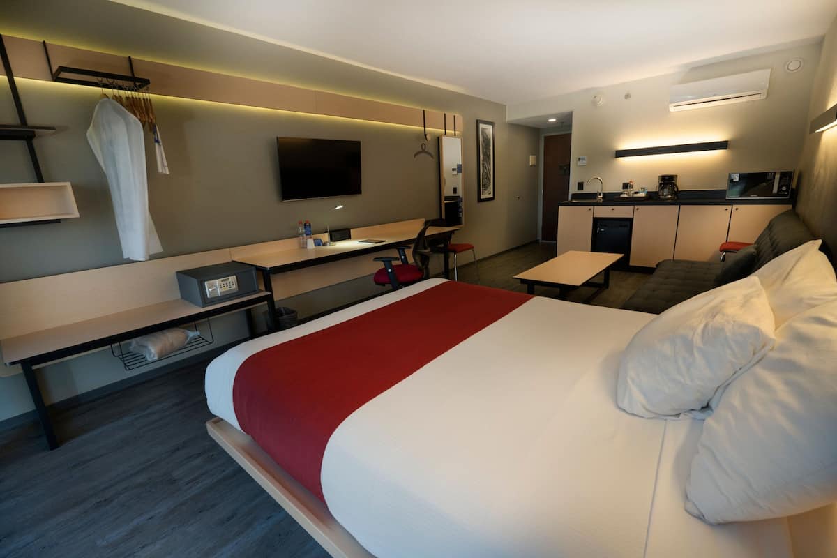 A modern hotel room with a large bed, a desk, a chair, a sofa, a TV, and a kitchenette.