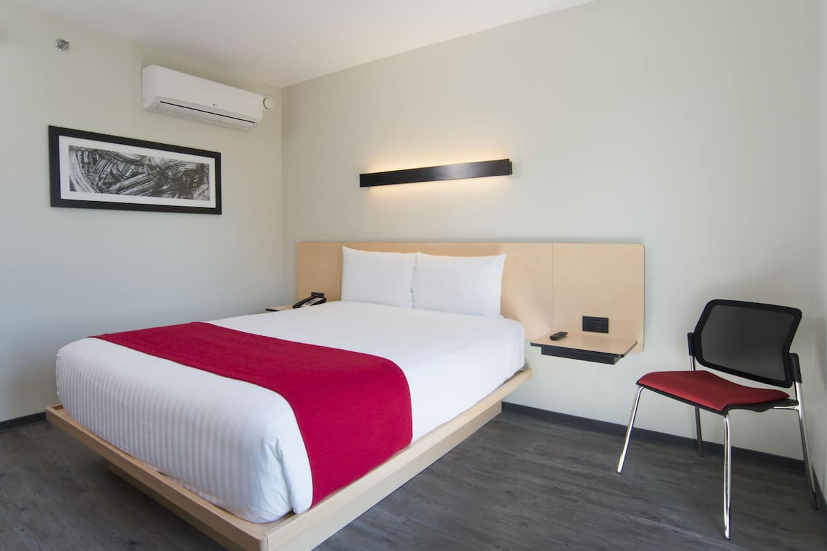 A hotel room with a bed, a chair, a bedside table, a wall-mounted air conditioner, a framed picture, and a wall-mounted light fixture.