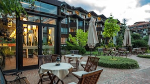 An outdoor patio area with tables and chairs, surrounded by greenery and a building with balconies. - Perun Lodge Hotel (Bansko)