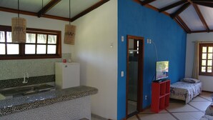 A kitchen with a tiled backsplash, a sink, and a countertop. There is a window with a view of trees, a door leading to another room, and a bed with a floral bedspread. - Pousada Capitão Gancho (Maraú)
