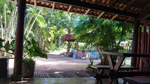 A covered outdoor area with wooden tables and chairs, surrounded by lush tropical plants and a thatched-roof structure. - Pousada Capitão Gancho (Maraú)