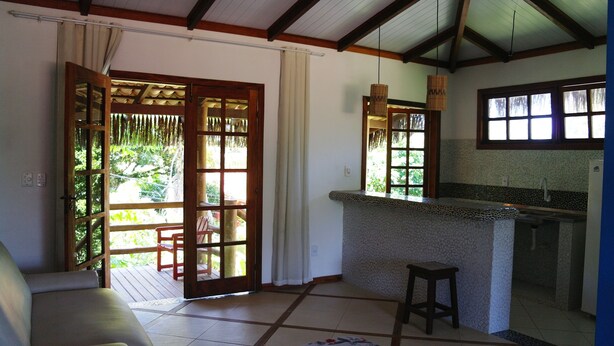 A room with a wooden ceiling, a kitchen area with a stone countertop, a sliding glass door leading to an outdoor deck, and a cushioned chair. - Pousada Capitão Gancho (Maraú)