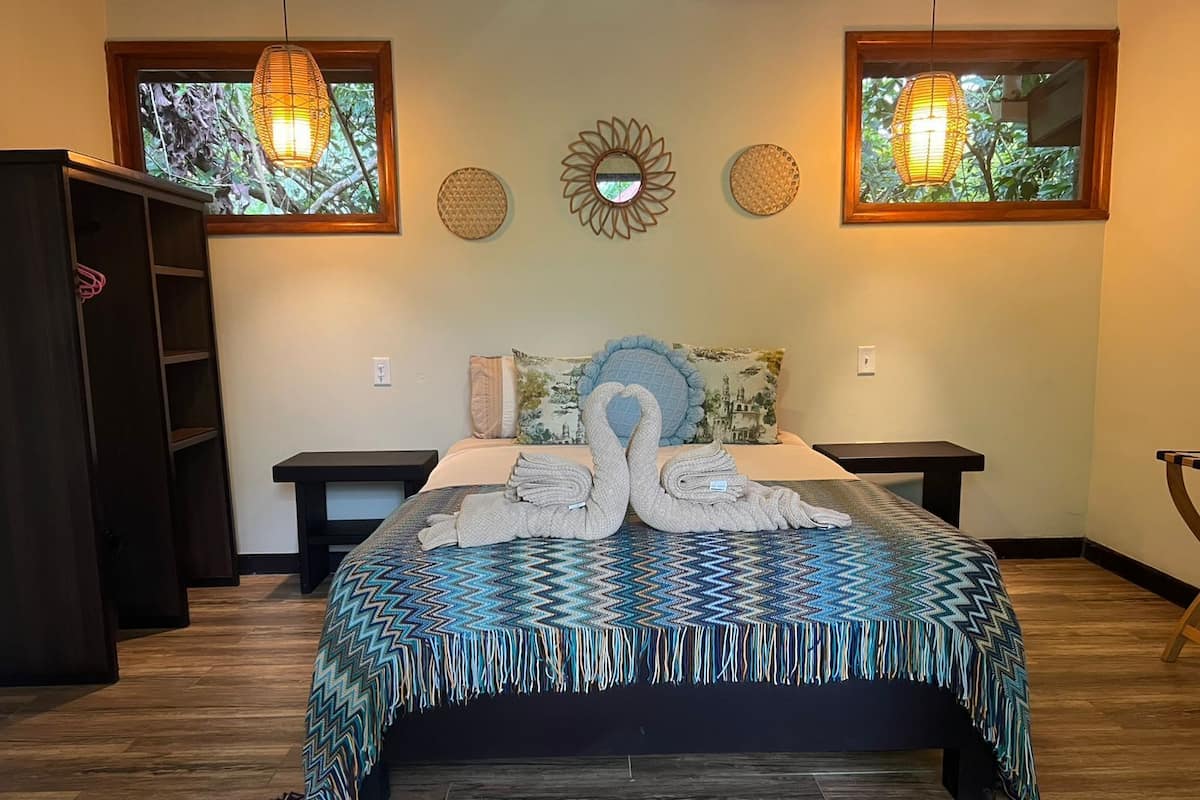 Una cama bien hecha con una colcha azul y verde estampada, dos lámparas fijadas a la pared y un espejo decorativo sobre la cama.