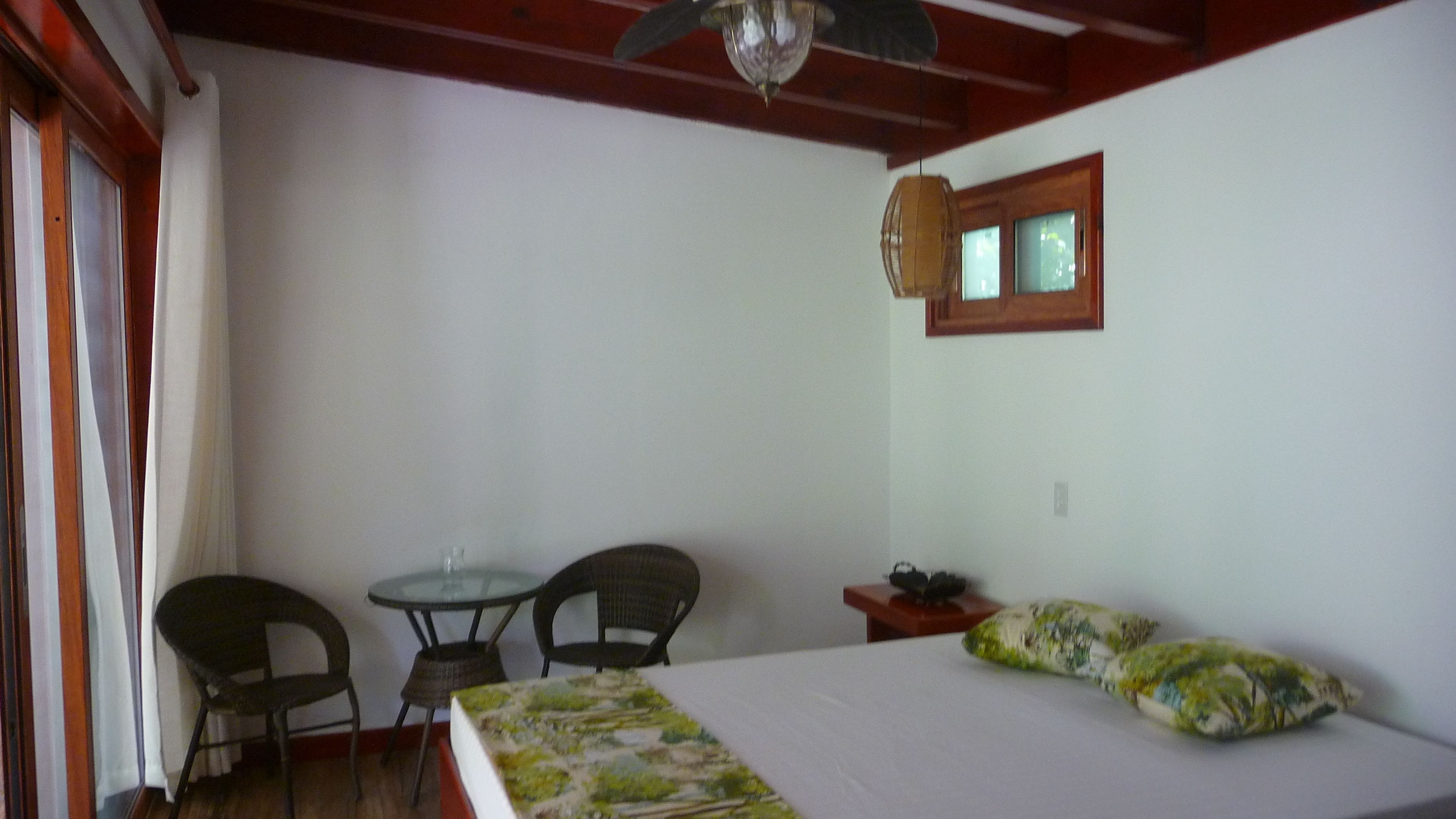 Una habitación con una cama, dos sillas, una mesita, un ventanal y un ventilador de techo.