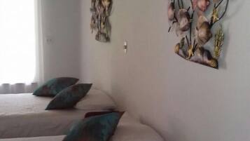 A hotel room with two beds, each with a white bedspread and teal pillows. There is a white standing fan on the right side of the room.