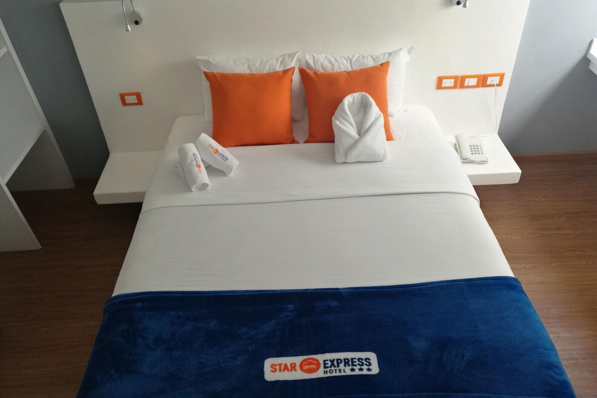 A hotel room with a bed, white bedding, orange pillows, a blue blanket, a phone, and a towel.