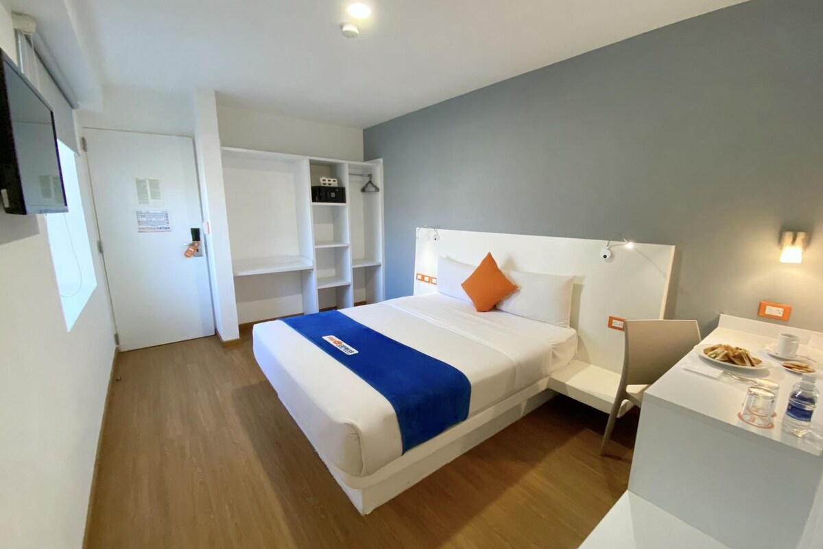 A hotel room with a bed, a desk, a chair, and a small kitchenette area.