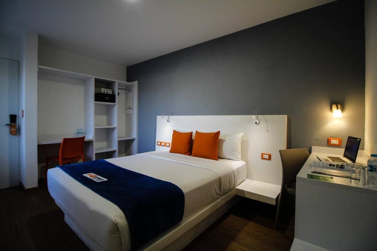 A hotel room with a bed, a desk, a chair, and a small workspace with a laptop.