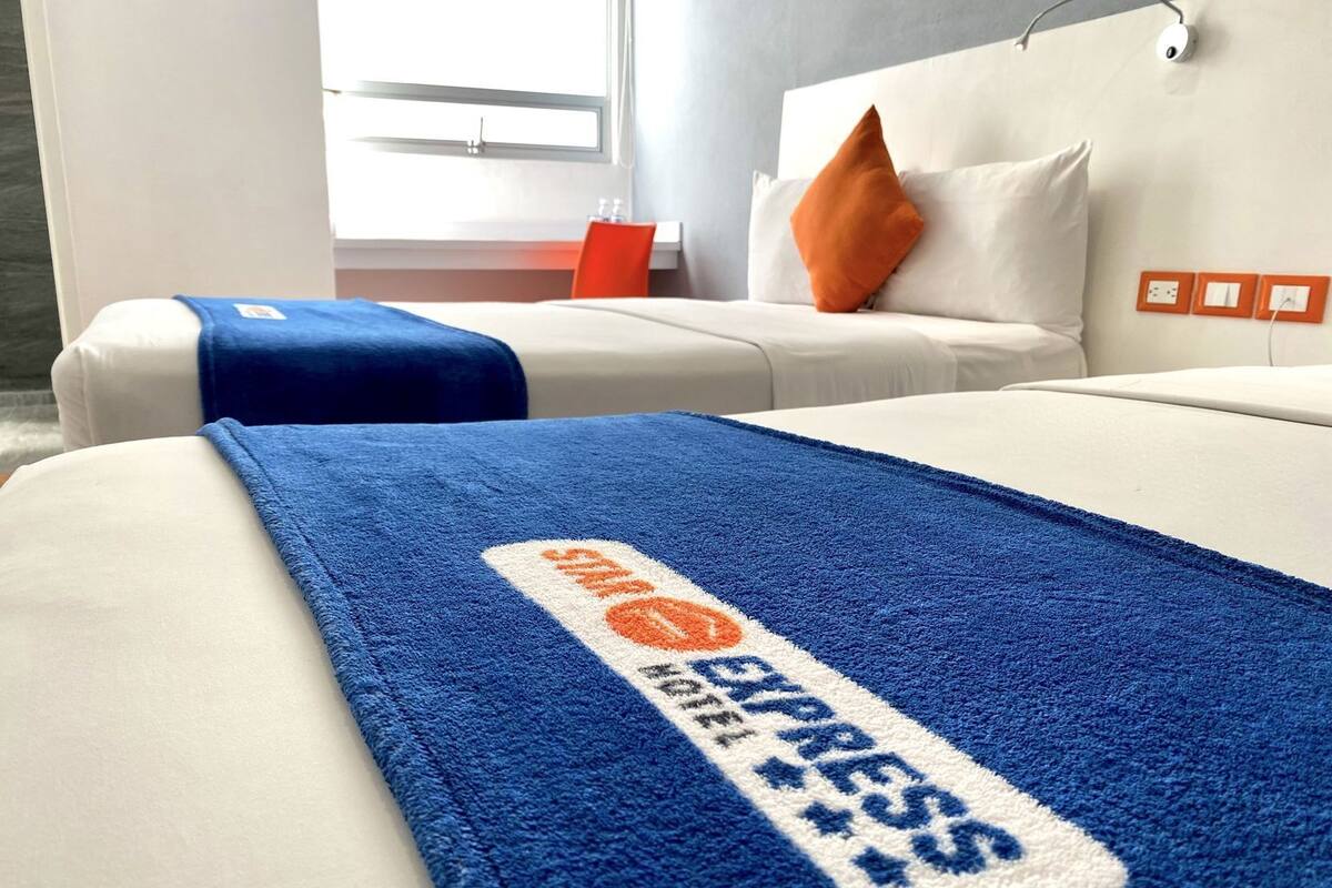 A hotel room with a bed, a blue blanket with a logo, a white headboard, and a window with daylight.