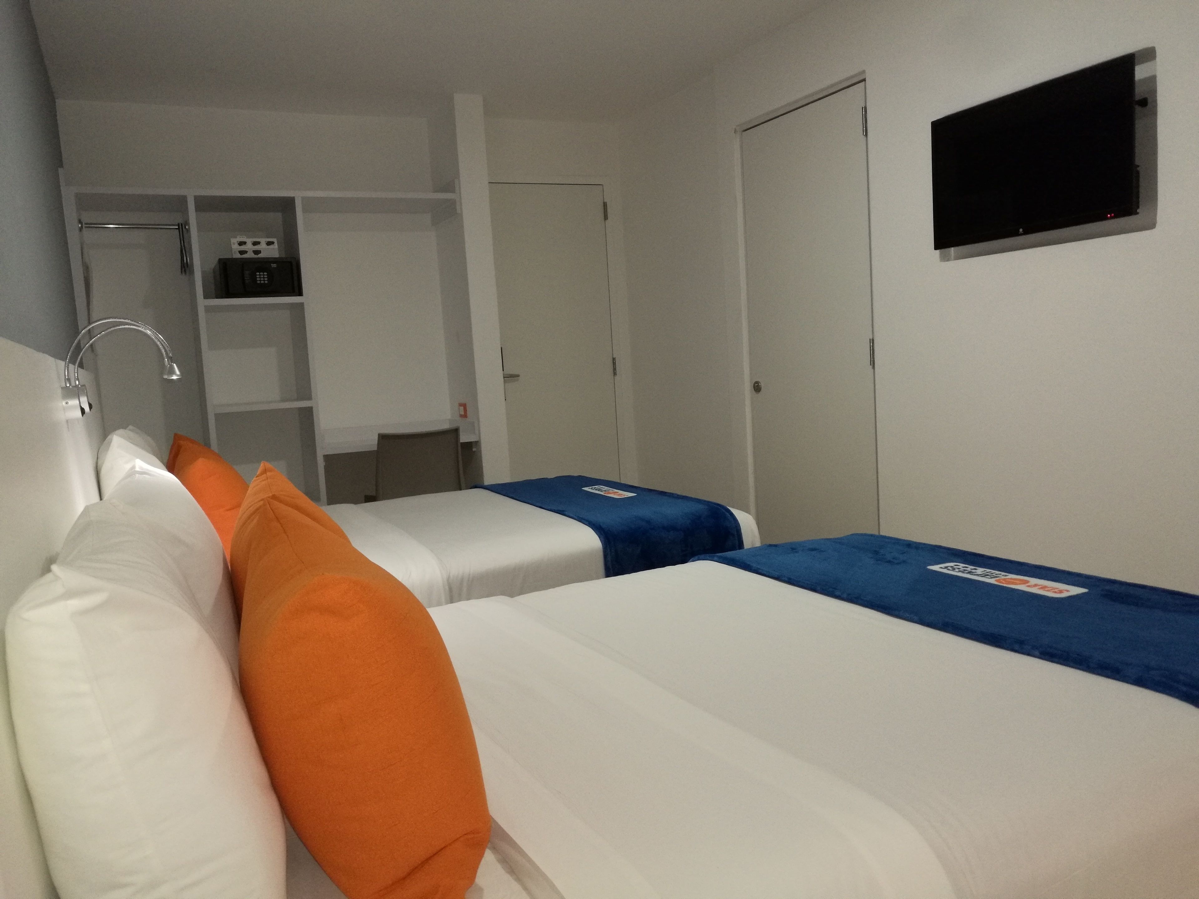 A hotel room with two beds, a TV mounted on the wall, a small kitchenette, and a wardrobe.